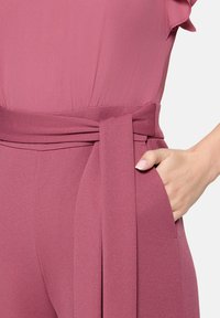 Rosa Overall mit einem Wickelbund, kurzen Rüschenärmeln und einer Seitentasche. Der Stoff wirkt glatt und leicht strukturiert.