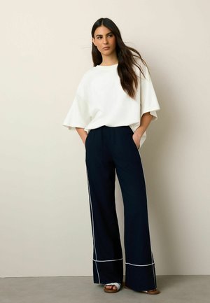 Woman with long dark hair wearing a loose white top, wide-leg navy pants with white piping, and white sandals, standing against a beige wall.
