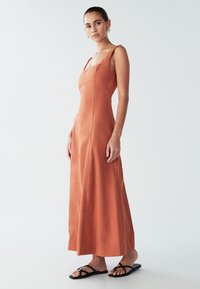 Maxi šaty farby hrdzavej s hranatým výstrihom, priliehavým živôtikom a splývavou sukňou. Vyrobené z hladkej látky, doplnené čiernymi plochými sandálmi.