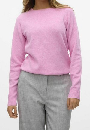 Kvinde iført en lyserød langærmet sweater og lysegrå skræddersyede bukser, stående mod en hvid baggrund.
