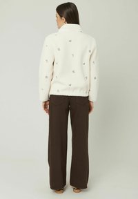 Veste en polaire écrue avec broderies florales, design avec col et finition texturée, associée à un pantalon large marron.