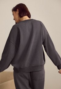 Šedá oversized mikina vyrobená z měkké tkaniny, s žebrovanými manžetami a lemem. Design na zadní straně má jednoduchý, bezšvový vzhled.