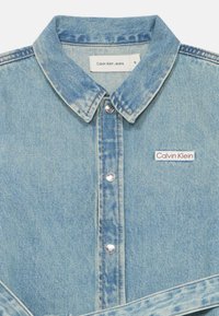 Svijetloplena traper košulja s klasičnom kragnom, srebrnim gumbima i crveno-bijelim Calvin Klein logom na lijevoj strani prsiju.