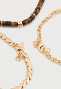 Deux bracelets chaîne en or avec fermoirs mousquetons et un bracelet de perles marron avec accents dorés sur un fond blanc.