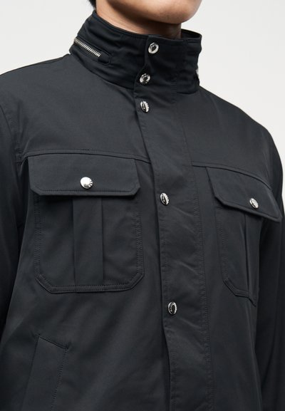 Κοντινό πλάνο προσώπου ατόμου που φοράει μαύρο μπουφάν με ψηλό γιακά, δύο τσέπες στο στήθος με ασημένια κουμπιά και κλείσιμο μπροστά με κουμπί πατιλέτα.