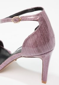 Chaussure à talon haut texturée violette avec bride à la cheville et boucle dorée, dotée d'un talon fermé et d'un design latéral ouvert.