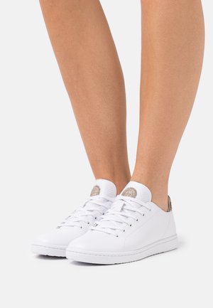 Nízké tenisky z bílé kůže se šňůrkami, nošené na nohou, s kulatou logo nášivkou na jazyku a strukturovanou patní záložkou.