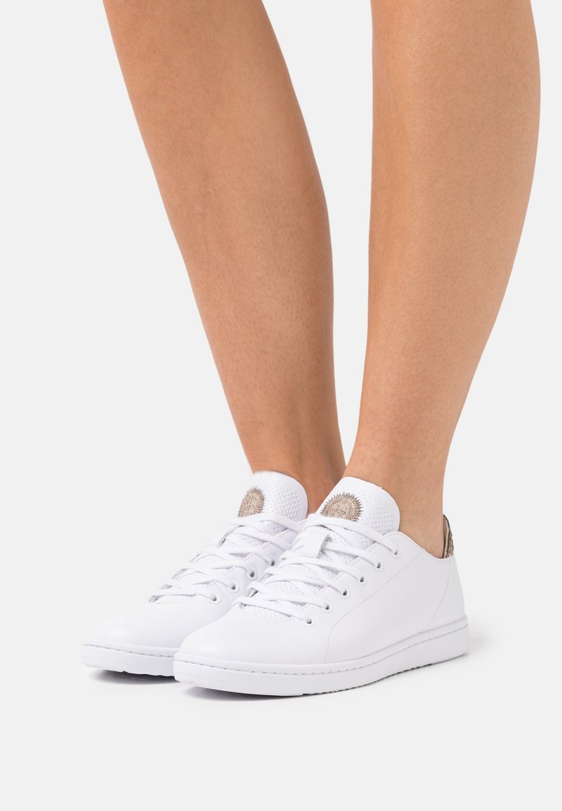 Baskets basses en cuir blanc avec lacets portées aux pieds, présentant un patch de logo rond sur la languette et un onglet de talon texturé.