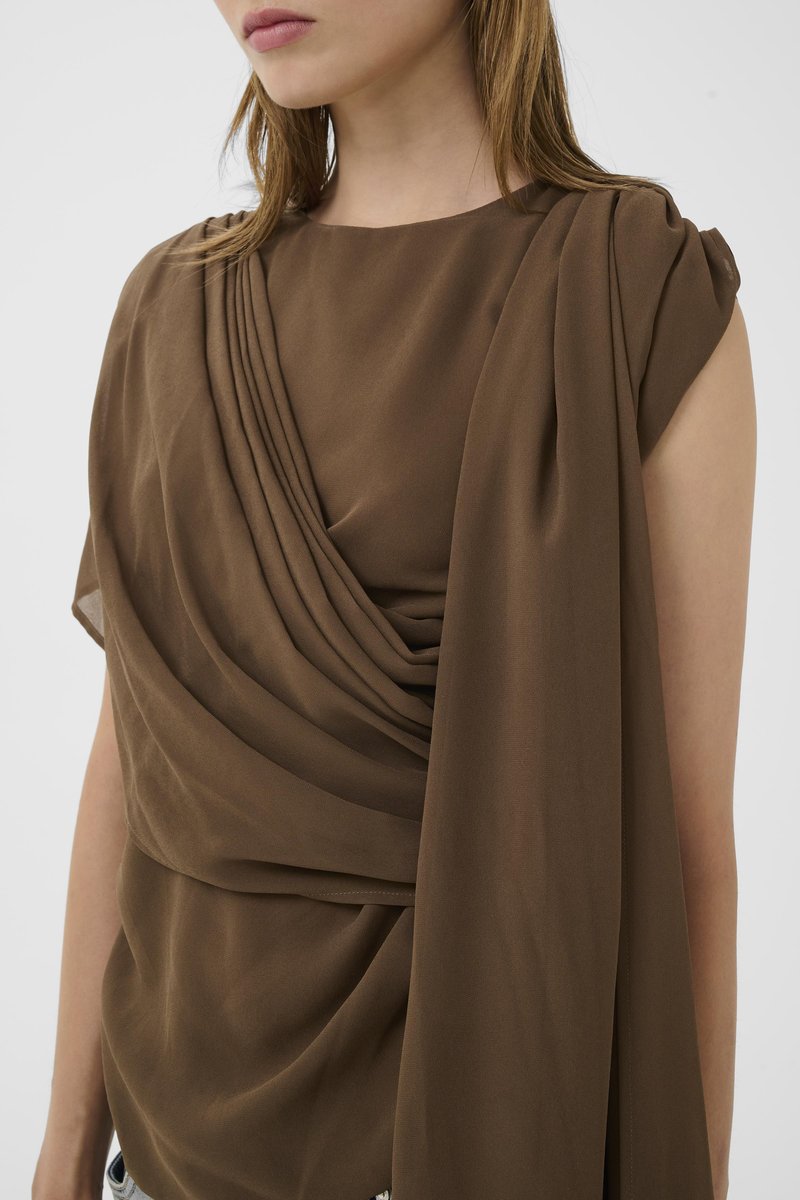 Femme portant un haut marron sans manches avec un tissu drapé et plissé sur une épaule sur un fond neutre.
