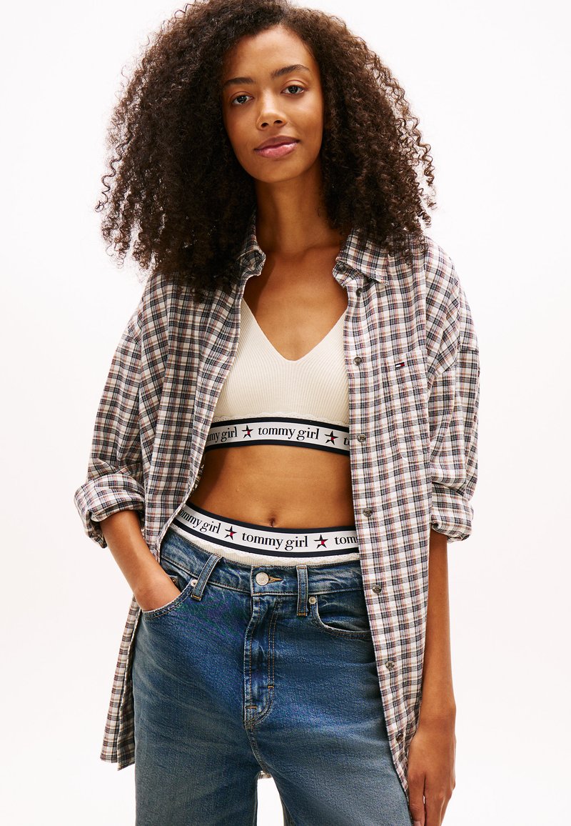 Camicia oversize a quadri grigia, blu e bianca, indossata sopra un top corto beige con fascia logo. Jeans blu a vita alta con tasche.