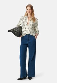 Camicia bianca con pois neri, design a bottoni, abbinata a jeans blu a vita alta e gamba larga e tacchi neri a punta, mentre tiene una borsa nera.