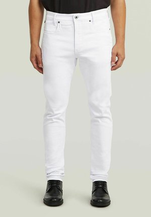 Homme portant un jean blanc slim avec cinq poches et des chaussures en cuir noir à lacets, debout devant un fond neutre.