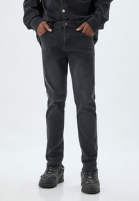 Jeans de mezclilla gris oscuro con corte ajustado, que cuentan con bolsillos frontales y un cierre de botón en la cintura, combinados con zapatillas negras texturizadas.