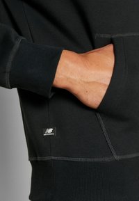 Black sweatshirt with a ribbed cuff and hem, featuring a front pocket. Includes a small “New Balance” logo label. Soft, textured fabric.