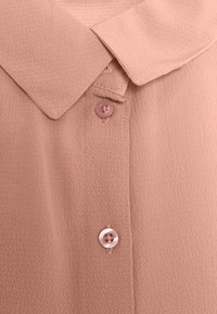 Blusa rosa chiaro in tessuto testurizzato con colletto a punta e due bottoni sul davanti. Liscia con una leggera lucentezza.