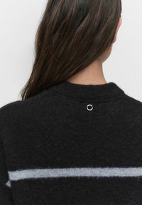 Svart ulltröja med hög halsringning, som har ett ljusgrått horisontellt rand och en silverfärgad cirkulär detalj i nacken.