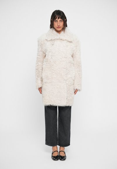 Cappotto in pelliccia sintetica bianca con colletto, dotato di maniche lunghe e silhouette dritta, abbinato a jeans grigi a gamba larga e scarpe nere.