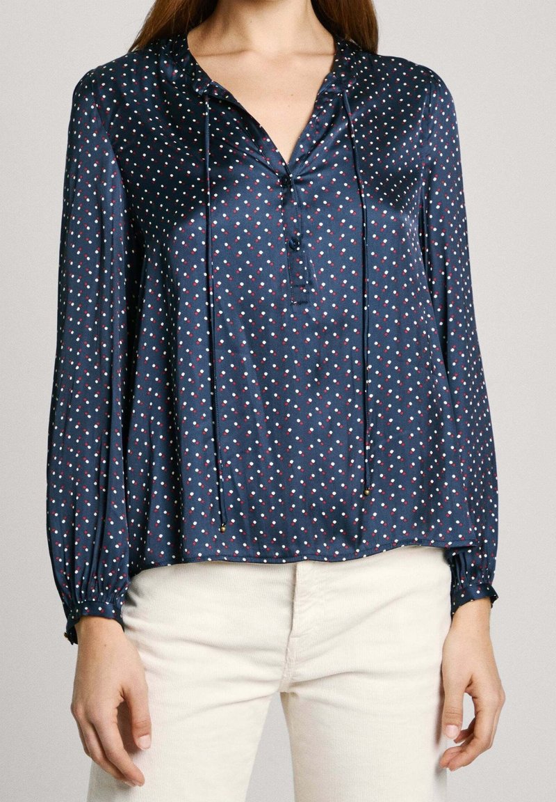 Chemise en satin bleu foncé à petits pois rouges et blancs, manches longues, devant boutonné, avec un design doux et drapé.