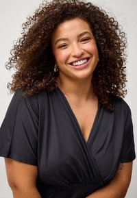 Haut enveloppant noir à manches courtes avec un décolleté en V, présentant une taille froncée et une texture de tissu douce. Personne souriante avec des cheveux bouclés.