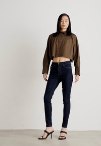 Smeđa skraćena košulja s dugmadi, uparena s tamnoplavim uskim trapericama. Model nosi crne sandale s petom, ističući elegantnu siluetu.