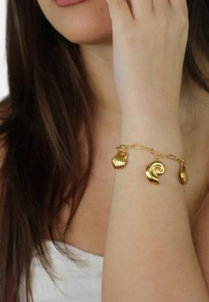 Mujer con cabello largo y castaño que lleva una pulsera de charms dorada con colgantes de concha y espiral en la muñeca izquierda, tocándose el rostro.