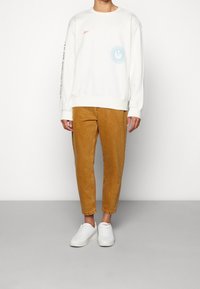 Felpa bianca con stampa grafica blu e logo arancione; abbinata a pantaloni tapered beige e sneakers bianche; texture complessiva liscia.