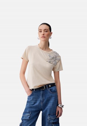 Donna con capelli scuri raccolti indossa una maglietta beige con decorazione floreale argento sulla spalla e jeans cargo blu con cintura nera.
