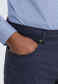Pantalons en coton bleu marine avec une texture lisse, dotés d'une poche avant et d'une fermeture à bouton en métal. Manches d'une chemise rayée bleue et blanche visibles.