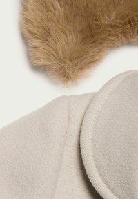 Beige uldstof med fiskebensmønster, der har en rund krave og et fluffy brunt pelsdetalje i øverste højre hjørne.