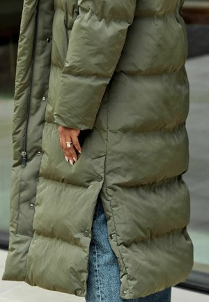 Olivově zelený péřový kabát s prošívaným designem. Přední zip, patky na knoflíky na lemu a boční rozparek. Nosený přes modré džíny.