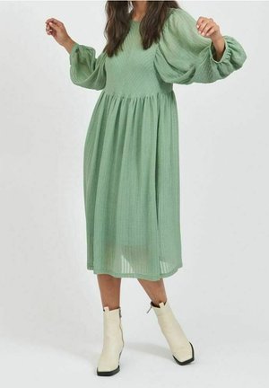 Robe verte arrivant au genou avec des manches bouffantes, un tissu texturé et une taille froncée. Portée avec des bottes blanches à talon moyen à bout carré.