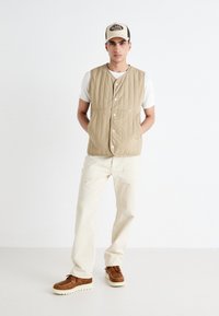 Chaleco beige acolchado con botones, llevado sobre una camisa blanca de canalé. Un par de pantalones beige claro y zapatos marrones con cordones y suela blanca.