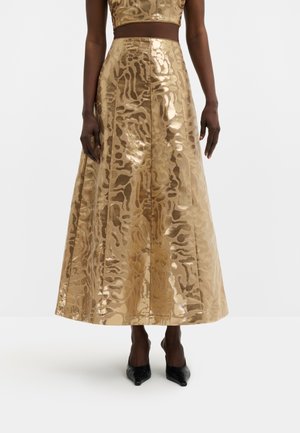 Femme portant une jupe midi dorée brillante à motifs avec une taille haute, associée à des talons noirs pointus, debout sur fond blanc.