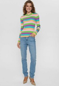 Donna sorridente, che indossa un maglione a righe colorato con maniche lunghe, jeans azzurri chiari e décolleté beige a punta, in piedi su uno sfondo semplice.