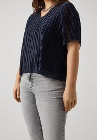 Mörkblå plisserad topp med V-ringning och korta ärmar, tillverkad av ett strukturerat tyg. Kombinerar med ljusgrå jeans för att visa enkelhet.