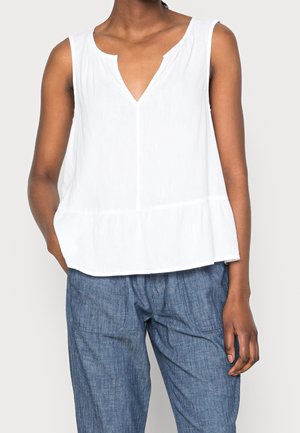 Mouwloze witte blouse met V-hals, gecombineerd met loszittende blauwe broek, gedragen door een persoon met één hand in de zak.