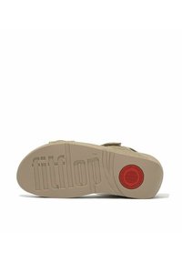 Sandalia beige con parte superior de tela texturizada, suela de goma con el logo "fitflop" y acento circular rojo cerca del talón. Diseño plano y de soporte.