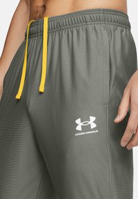 Šedé športové kraťasy s textúrovaným povrchom, s bielym logom Under Armour a žltou šnúrkou, ukazujúce priliehavý, zúžený strih.