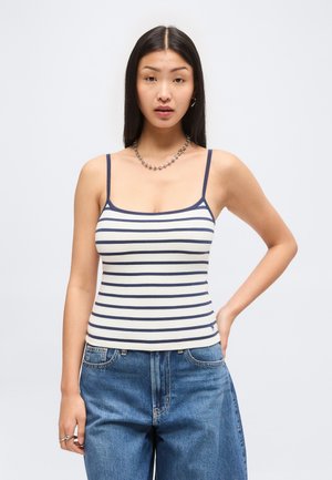 Young woman with long dark hair wearing a white and navy striped tank top and blue denim jeans, standing with one hand on her hip.