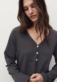 Maglietta a maniche lunghe grigio scuro con scollo a V, dotata di patta con bottoni e texture di tessuto morbido, indossata da un modello con capelli lunghi.