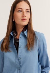 Hellblaue Jeansbluse mit durchgehender Knopfleiste, spitzem Kragen und Brusttasche, kombiniert mit einem braun-weißen gestreiften Schal.