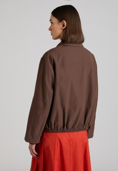 Donna con capelli castani lisci che indossa una giacca marrone con orlo elasticizzato e una gonna plissettata arancione, vista di spalle su uno sfondo semplice.