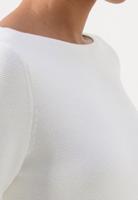Gros plan d'une personne portant un haut en maille texturée blanche avec un large col rond sur un fond uni.