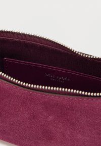 Pochette en daim bordeaux avec fermeture éclair argentée et poche intérieure présentant le logo "kate spade NEW YORK" embossé sur la doublure.