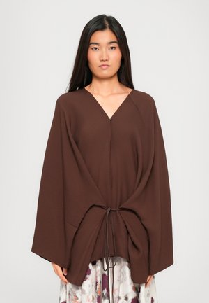 Woman with long black hair wearing a loose brown V-neck top with wide sleeves and a tie detail over a floral skirt.