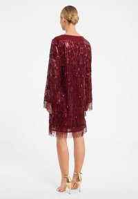 Bordeaux franjejurk met lange mouwen, verstevigd met pailletten, heeft een recht model en franjes aan de onderkant. Gouden hoge haksandalen.