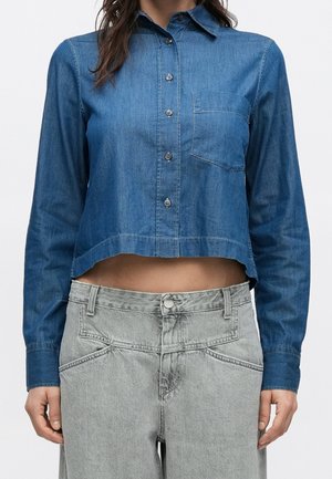 Femme portant une chemise en jean bleue courte à manches longues avec fermeture par boutons et un jean gris taille haute.