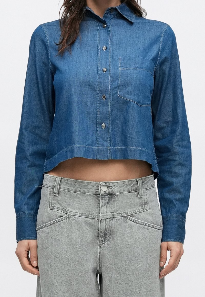 Femme portant une chemise en jean bleue courte à manches longues avec fermeture par boutons et un jean gris taille haute.