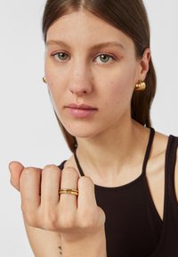 Złoty pierścionek o gładkim wykończeniu i smukłym kształcie, noszony na dłoni. Modelka ma na sobie pasujące złote kolczyki w kształcie kółek o zaokrąglonym wzorze.