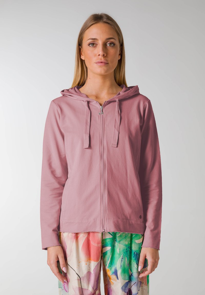Ljust rosa zip-up hoodie med dragsko på huvan, långa ärmar och avslappnad passform. Kombinerad med färgglada blommönstrade byxor.